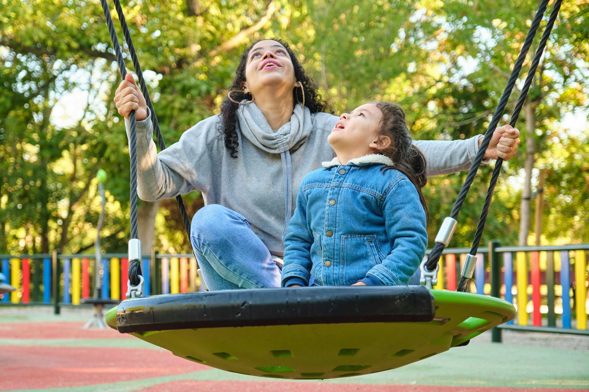 Happy Latin mother playing with her son looking up on a swing in a park.